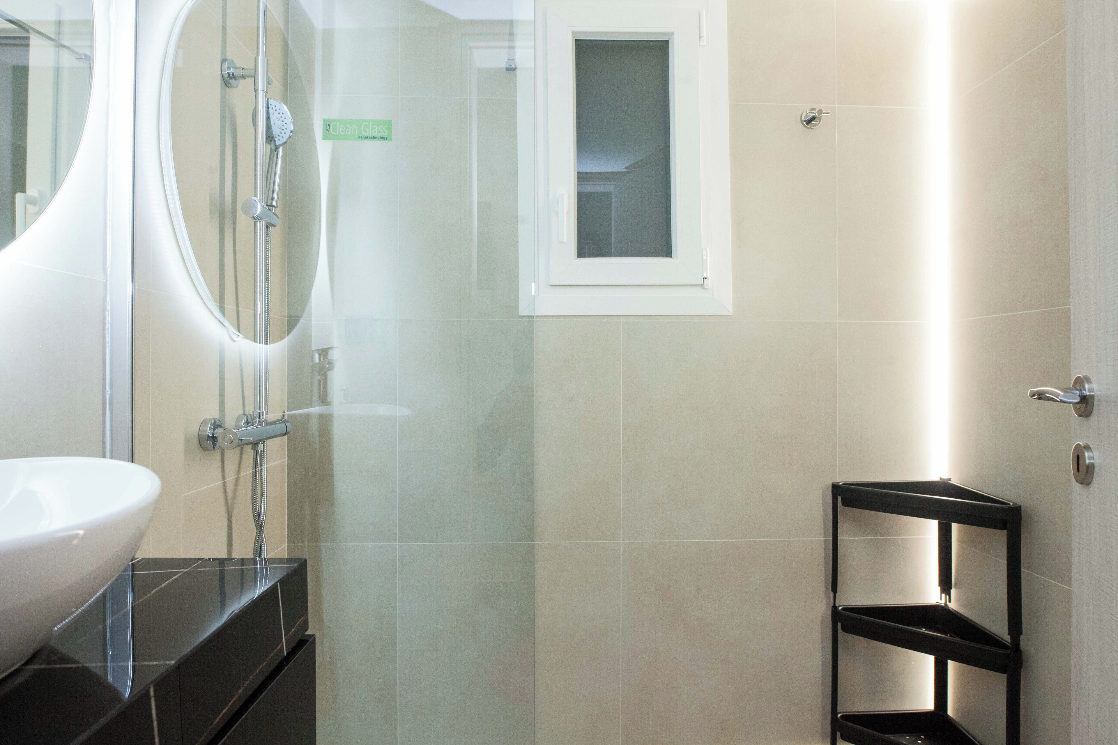 Un baño con ducha de vidrio, un lavamanos blanco y un estante negro.
