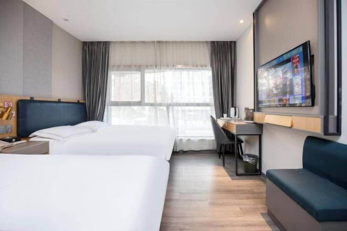 A hotel room with two beds, a desk, a chair, a TV, and a large window with curtains.