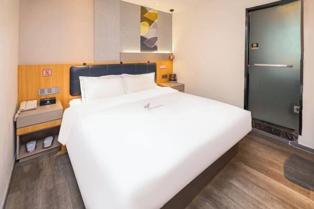 A hotel room with a large bed, a nightstand with a phone and a lamp, a wooden headboard, and a door with a handle.