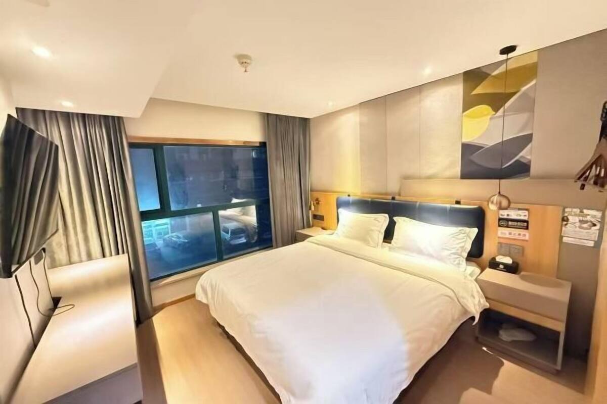 A modern hotel room with a large bed, a bedside table, a nightstand, a window with curtains, and a wall-mounted artwork.
