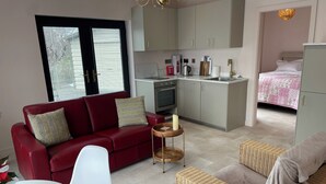 A compact living space with a red sofa, white chair, wicker ottoman, kitchenette, and a bed with a pink quilt. - Lakeside Apartment Retreat in Oughterard Connemara (Oughterard)