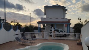 Una piscina con una terraza blanca, un área de patio cubierta y una camioneta blanca estacionada cerca.