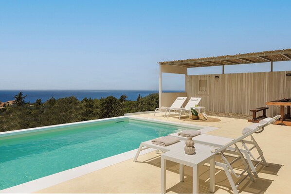 Área da piscina com espreguiçadeiras e mesa, com vista para o mar.