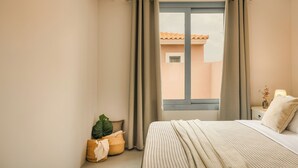 A bedroom with a bed, curtains, a nightstand, a lamp, and a view of buildings outside. - Villa Ivi - Zen Villas (Fiskardo)