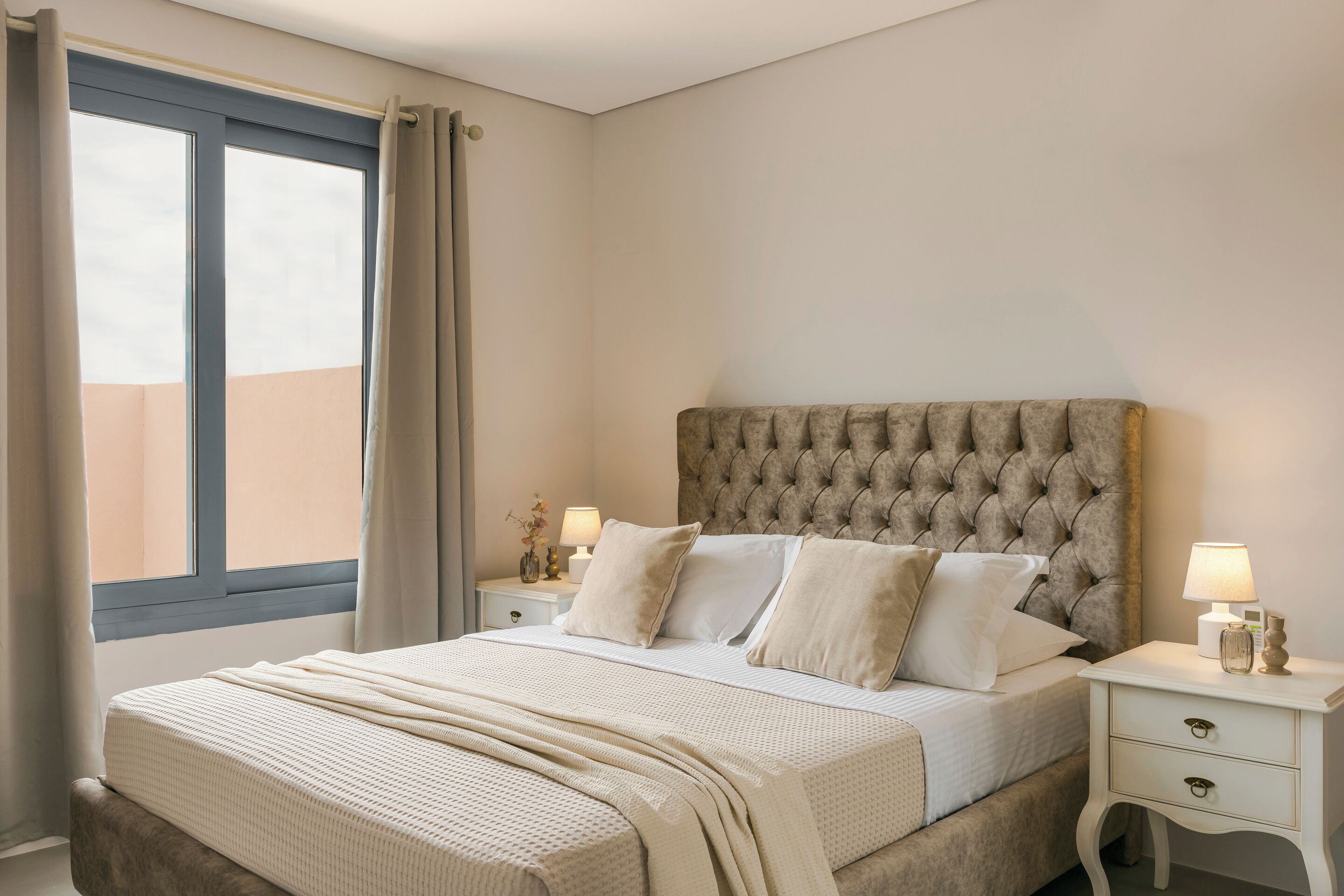 Una camera da letto con testiera trapuntata, un'ampia finestra con tende, un comodino con lampada e un letto con copriletto e cuscini bianchi.