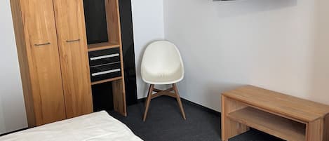 A hotel room with a bed, a wooden wardrobe, a TV, a white chair, and a wooden bench.