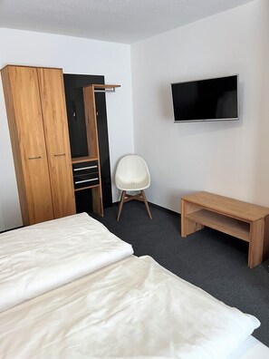 A hotel room with a bed, a wooden wardrobe, a TV, a white chair, and a wooden bench. - Altötting City Apartments (Altötting)