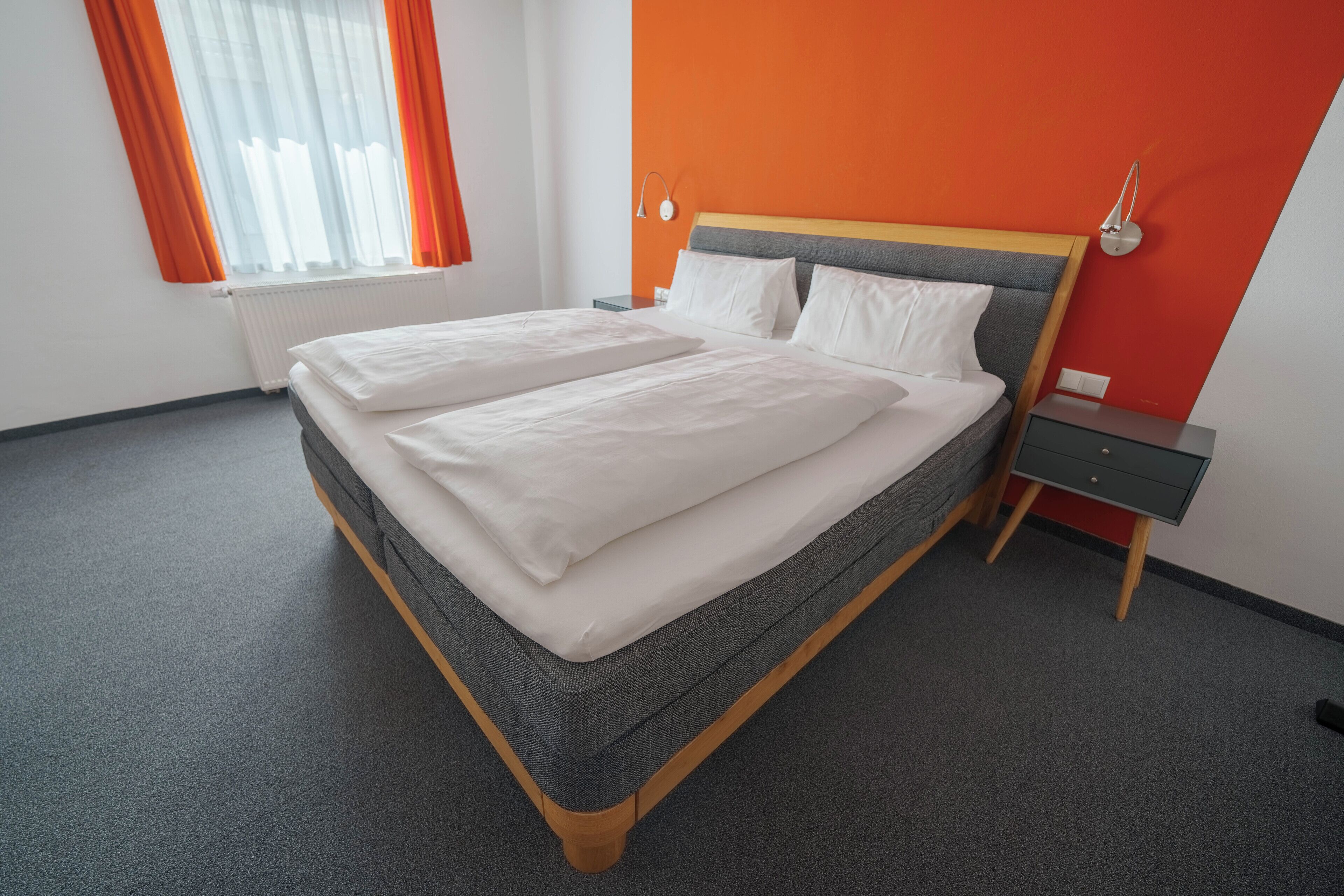 A neatly made double bed with white linens, a grey headboard, and a wooden frame. There is a small bedside table with a lamp and a power outlet on the wall.