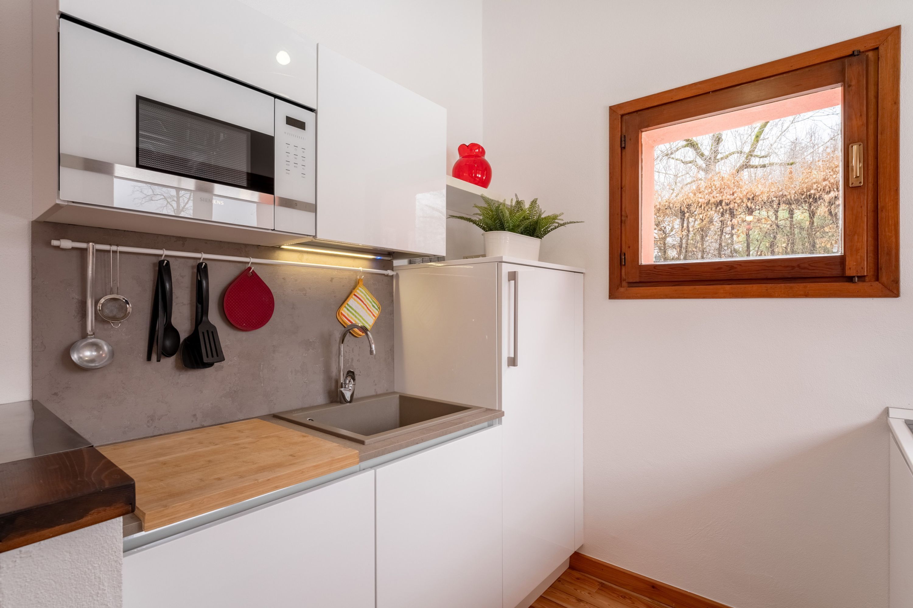 A modern kitchen with white cabinetry, a stainless steel sink, and a microwave. There is a window with a view of trees and a red decorative item on the wall.