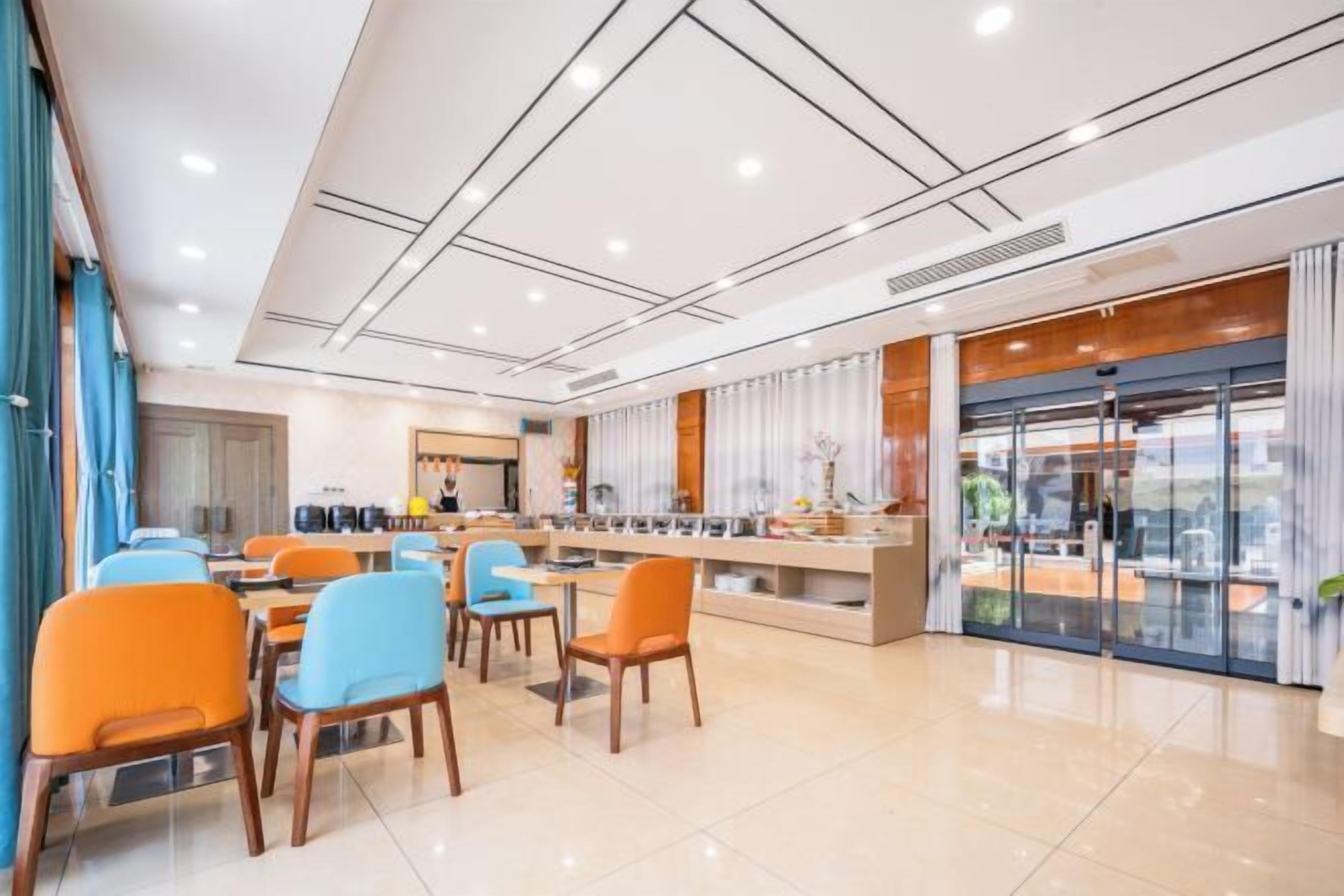 A modern dining area with multiple tables and chairs, a kitchen in the background, and a glass partition with curtains.