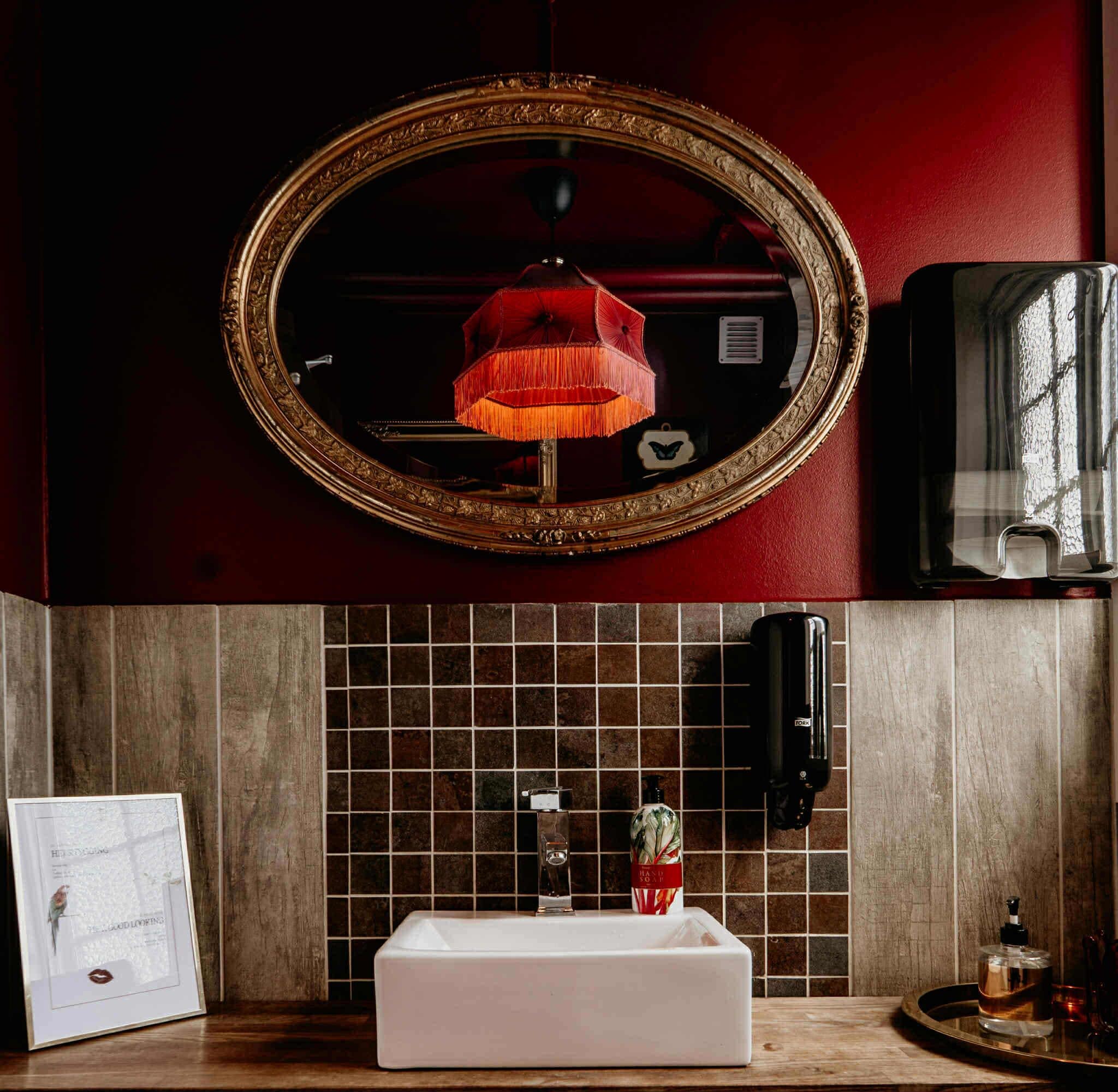 Un baño con un espejo grande, una lámpara colgante roja, un lavabo blanco, un dispensador de jabón y un secador de manos.
