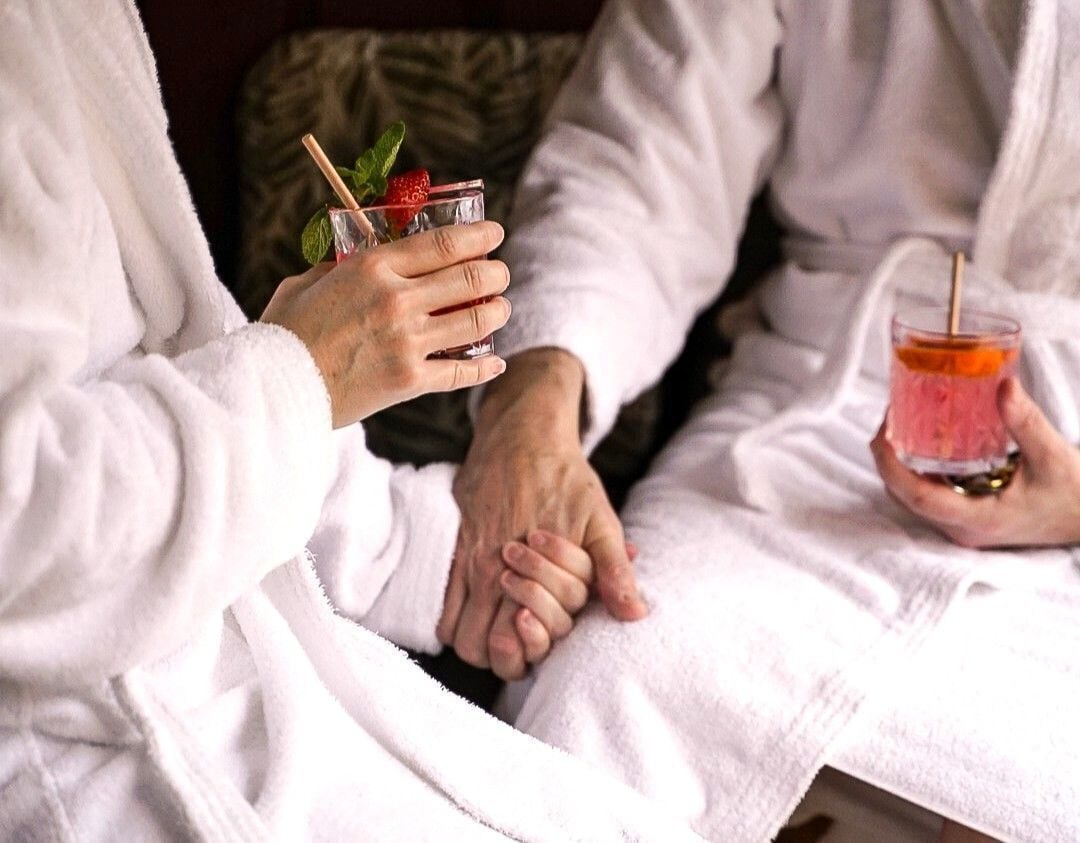 Dos personas vestidas de blanco sosteniendo bebidas con pajitas y adornos, sentadas muy cerca la una de la otra.