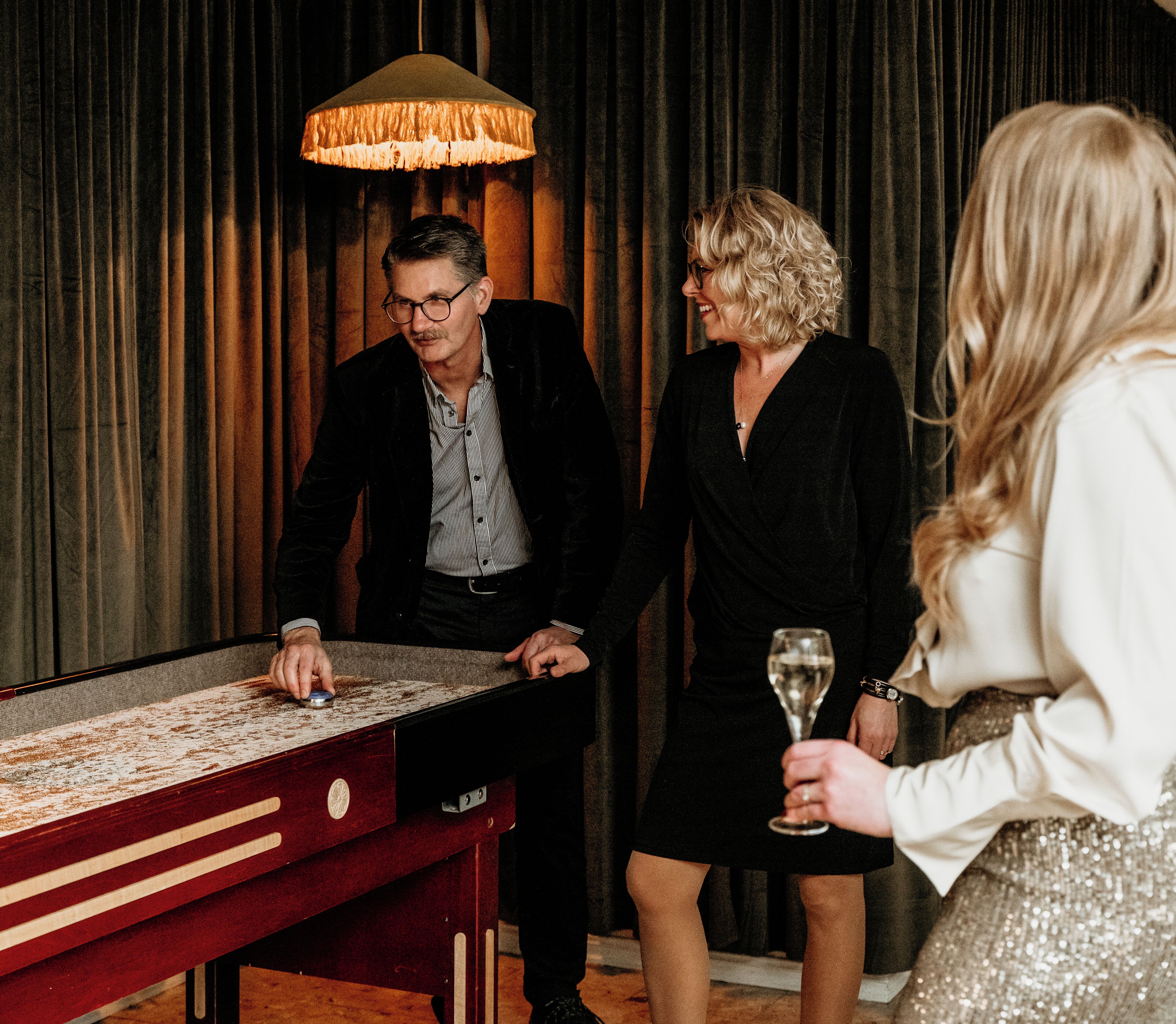A group of people around a table with a game, a lamp, and a curtain in the background.
