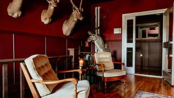 A room with wooden flooring, two upholstered chairs, a table with a candle, and mounted animal heads on the wall.