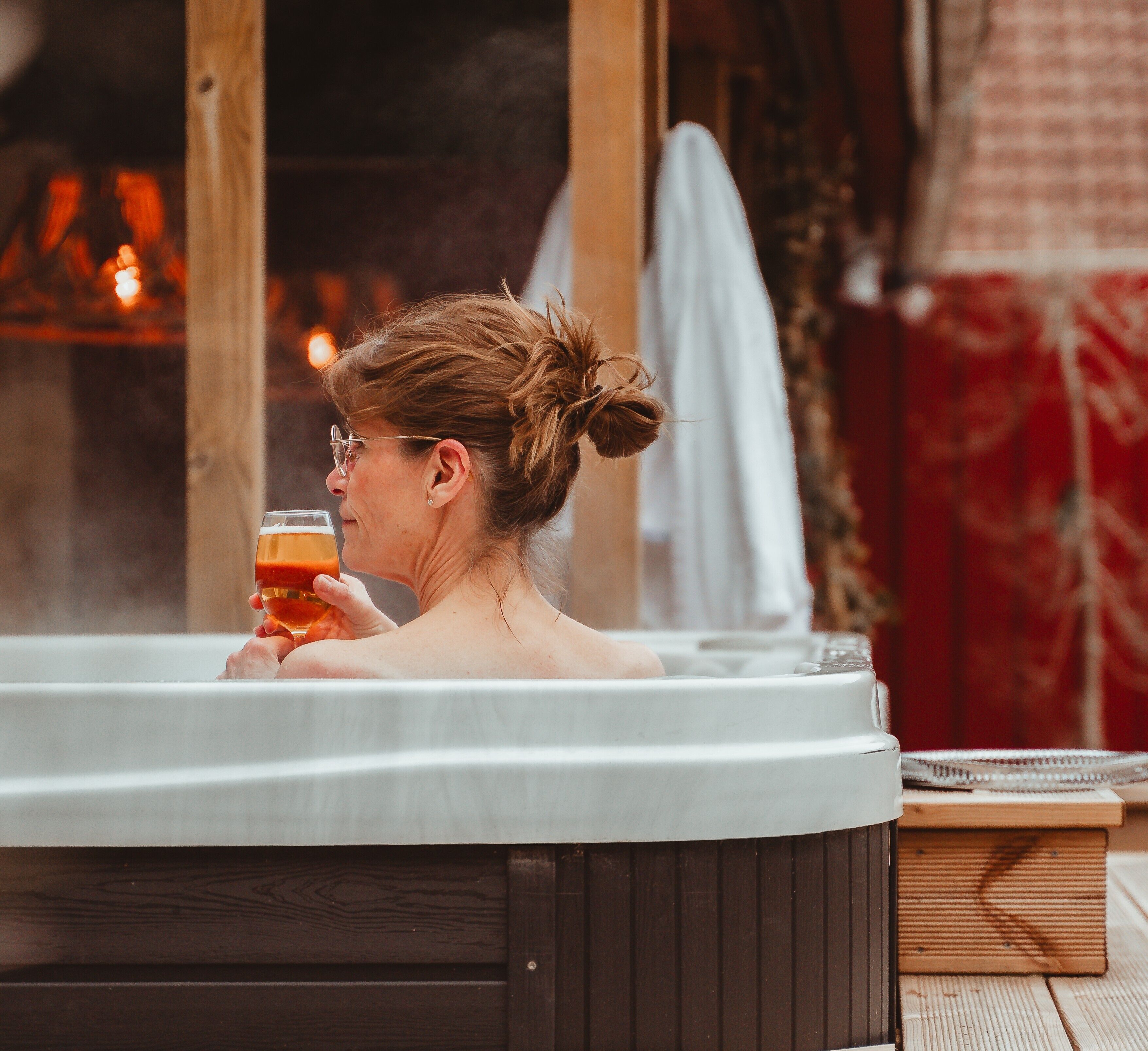 En person i ett bubbelbad med ett glas i handen, en trästuga i bakgrunden och en handduk.