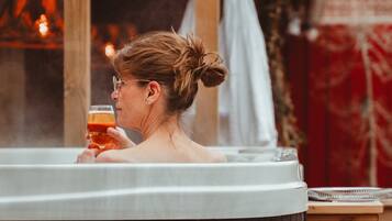 En person i ett bubbelbad med ett glas i handen, en trästuga i bakgrunden och en handduk.