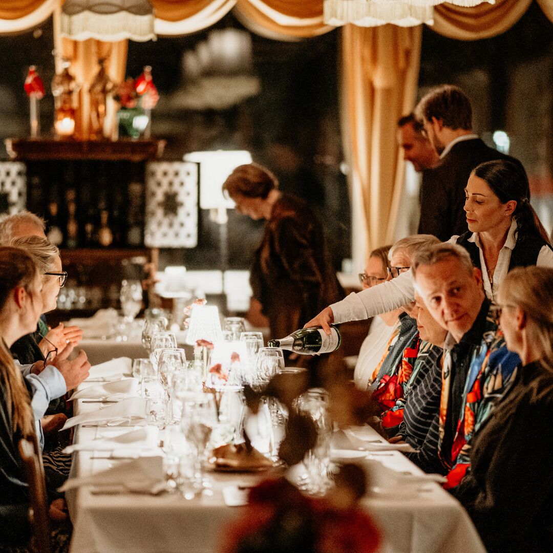 A group of people seated at a table with a white tablecloth, glasses, and a bottle being poured.
