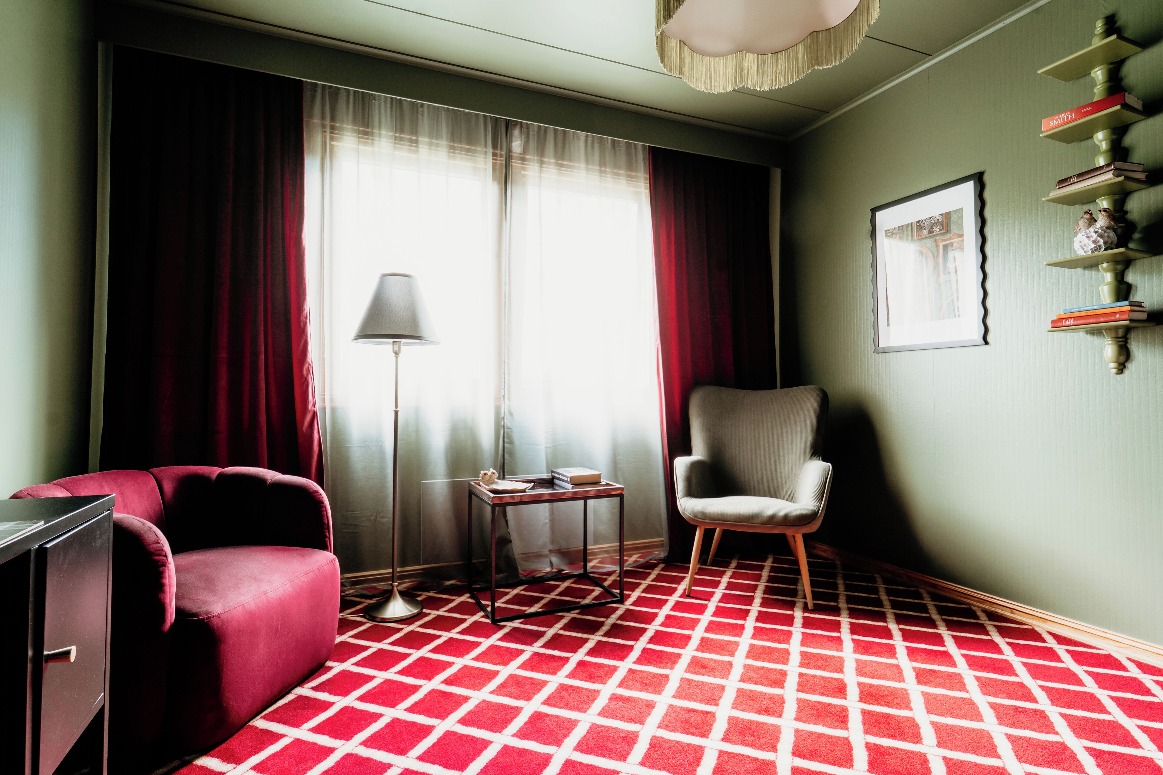 Habitación de hotel con alfombra a cuadros rojos y blancos, un sofá rojo, un sillón gris, una mesita con una lámpara y una estantería con libros.