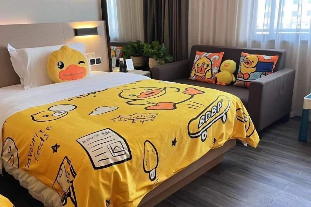 Hotellihuone, jossa on keltainen, Ankronikkaloita-aiheinen peitto sängyllä, harmaa sohva, jossa on Ankronikkaloita-aiheisia koristetyynyjä, ja ikkuna, jossa on verhot.