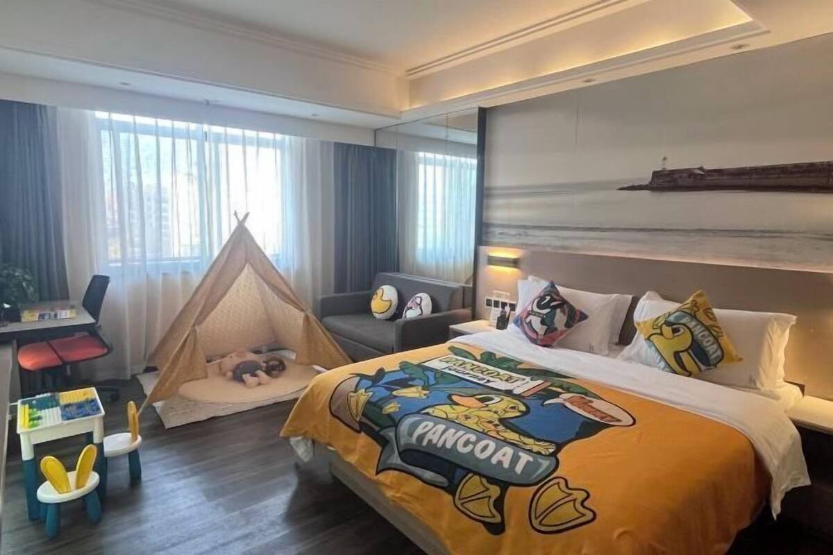 Hotellihuone, jossa on sänky, jonka päällä on «Pancooat»-teemalla koristeltu päiväpeite, työpöytä tuolilla, pieni leikkialue, jossa on teltta, ja suuret ikkunat, joista avautuu näkymä merelle.
