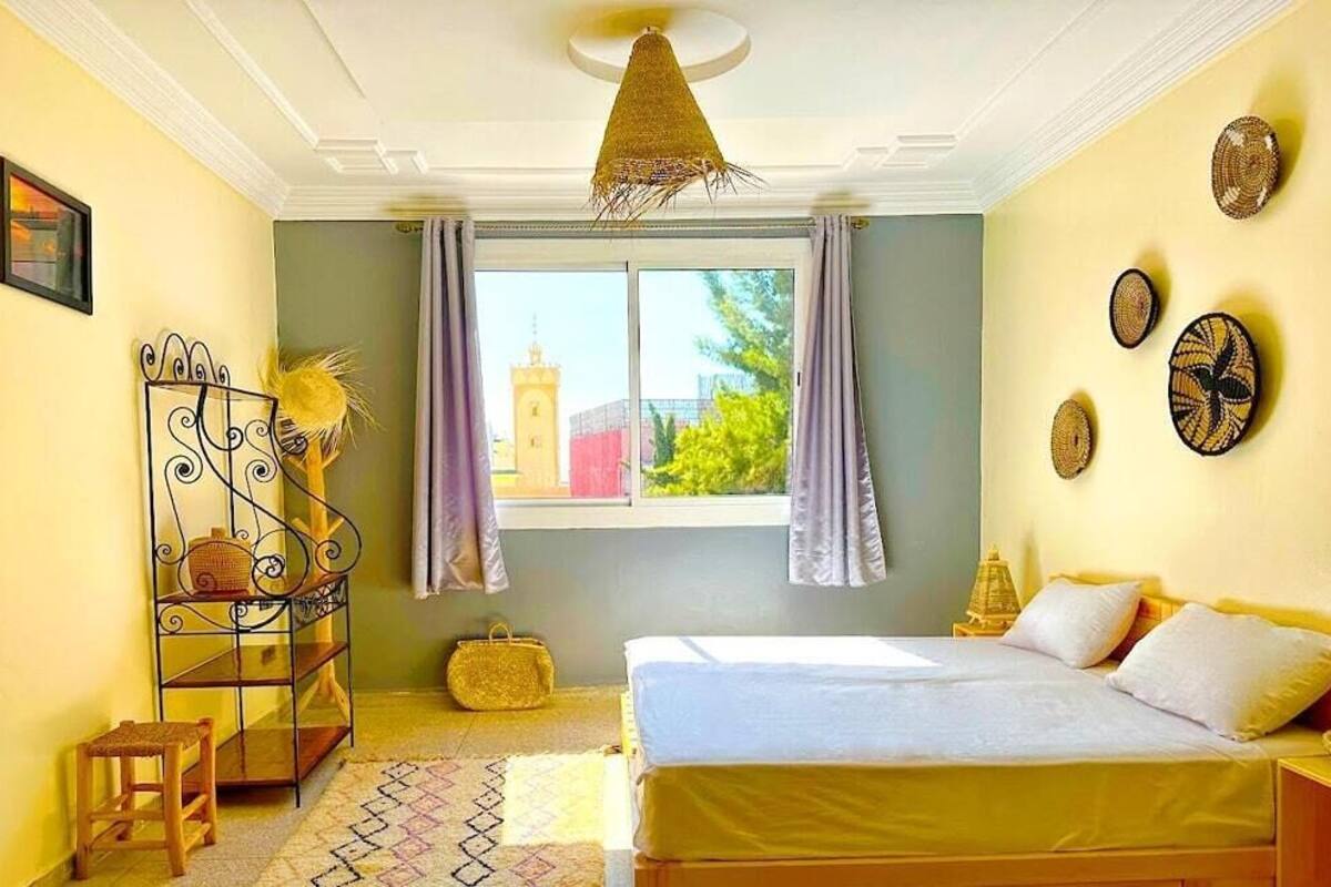 A bedroom with a bed, a window with curtains, a hanging light fixture, a small table with a chair, and decorative wall art.