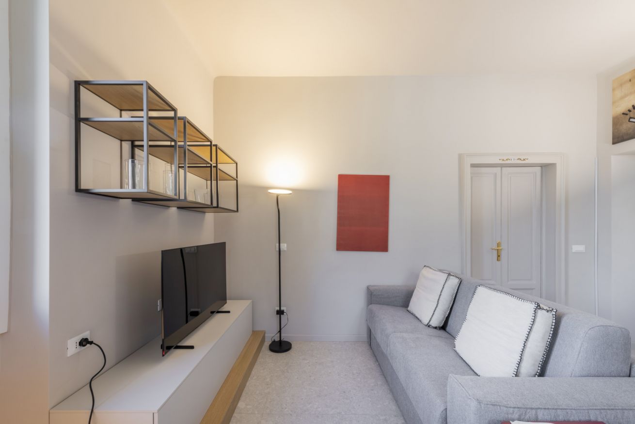 Uma sala de estar moderna com televisão de tela plana, abajur, sofá cinza com almofadas decorativas e uma parede de destaque vermelha.