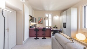 Una cocina compacta con una isla central, dos taburetes de bar, una zona de comedor con mesa y sillas, y un sofá en la sala de estar.