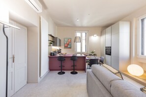 A compact kitchen with a central island, two bar stools, a dining area with a table and chairs, and a sofa in a living area.