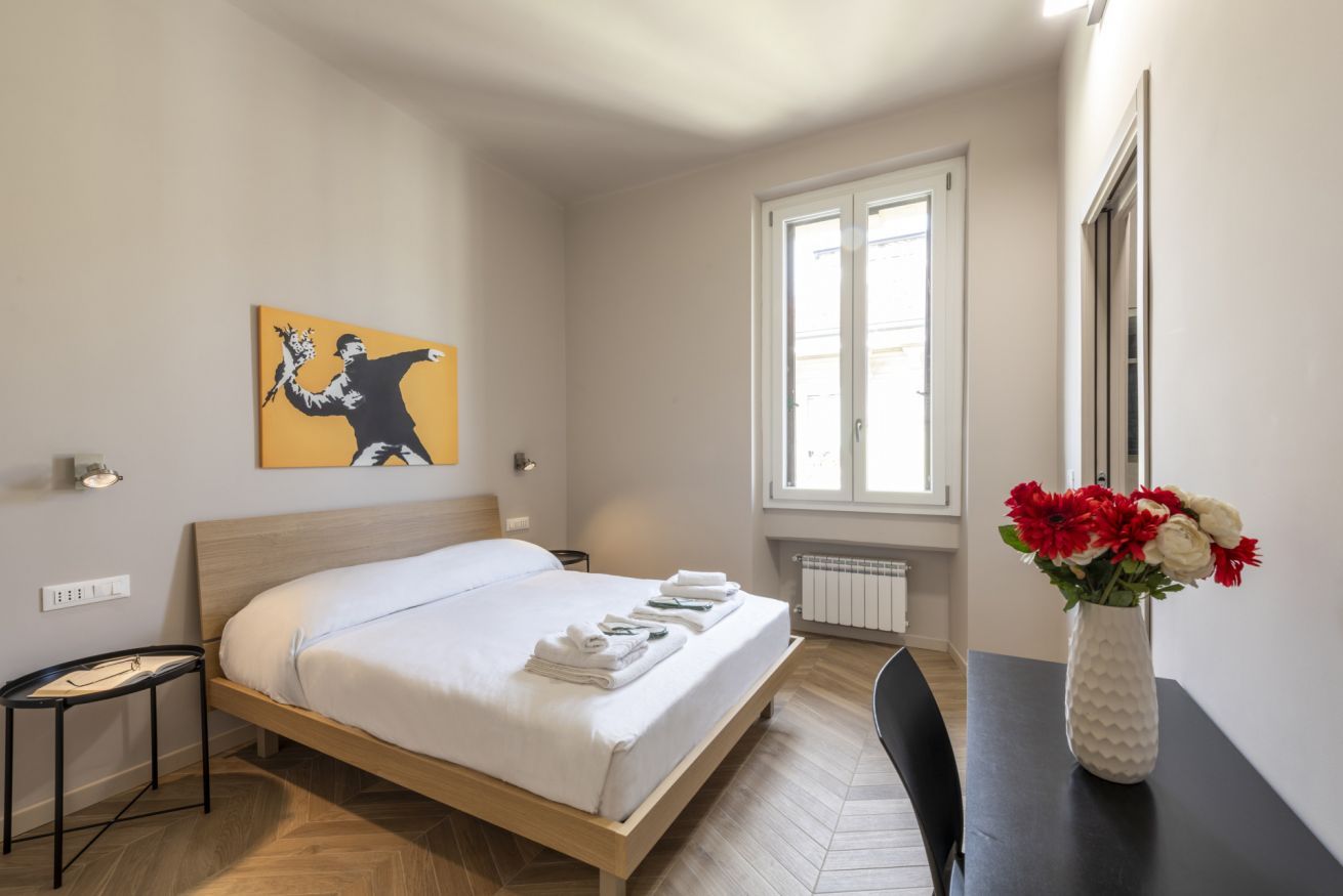 A hotel room with a bed, a window, a painting on the wall, and a vase with flowers on a table.