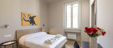 A hotel room with a bed, a window, a painting on the wall, and a vase with flowers on a table.