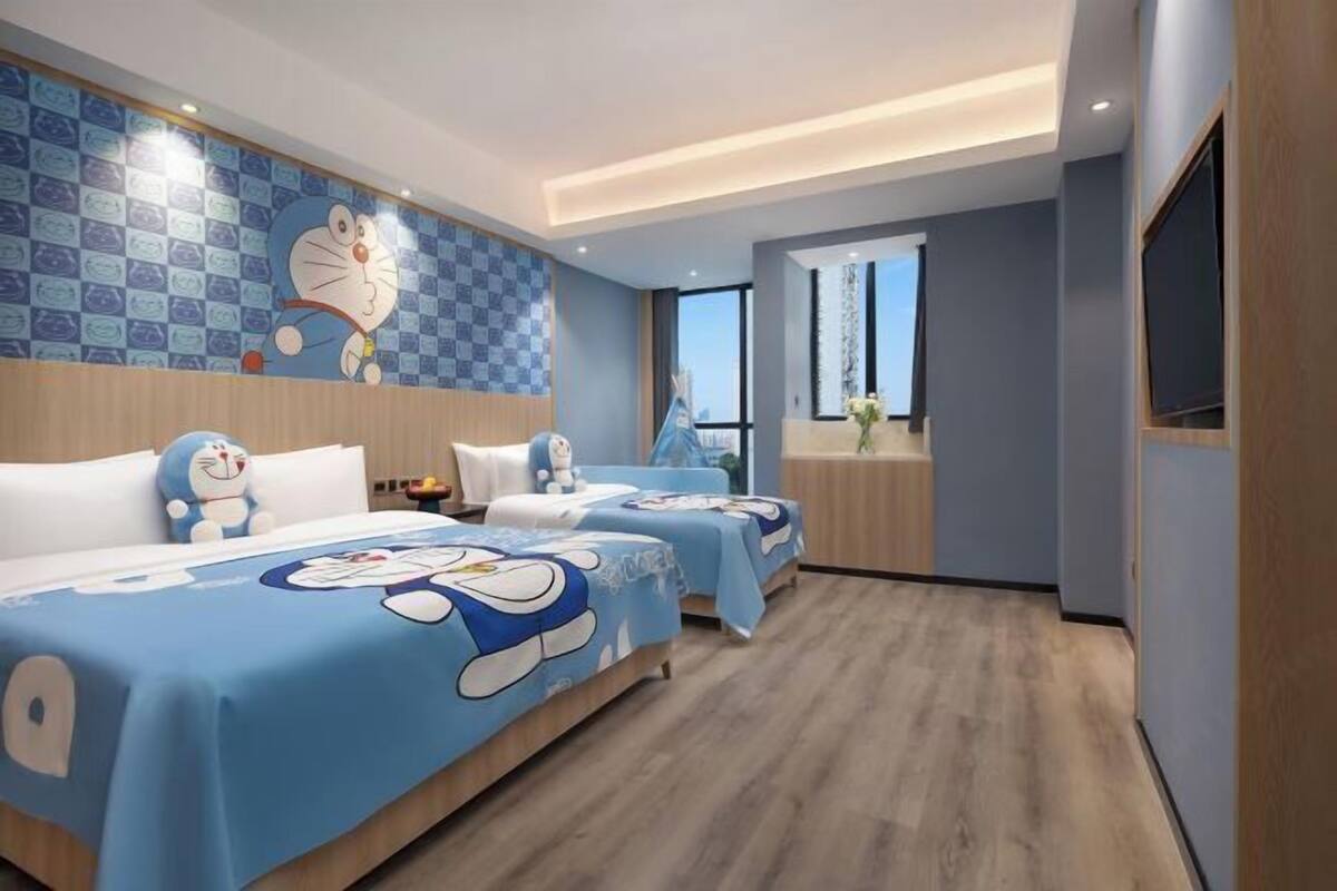 A hotel room with two beds, each covered with a blue blanket featuring cartoon characters. The room has a wooden floor, a large window, and a wall with a cartoon character mural.