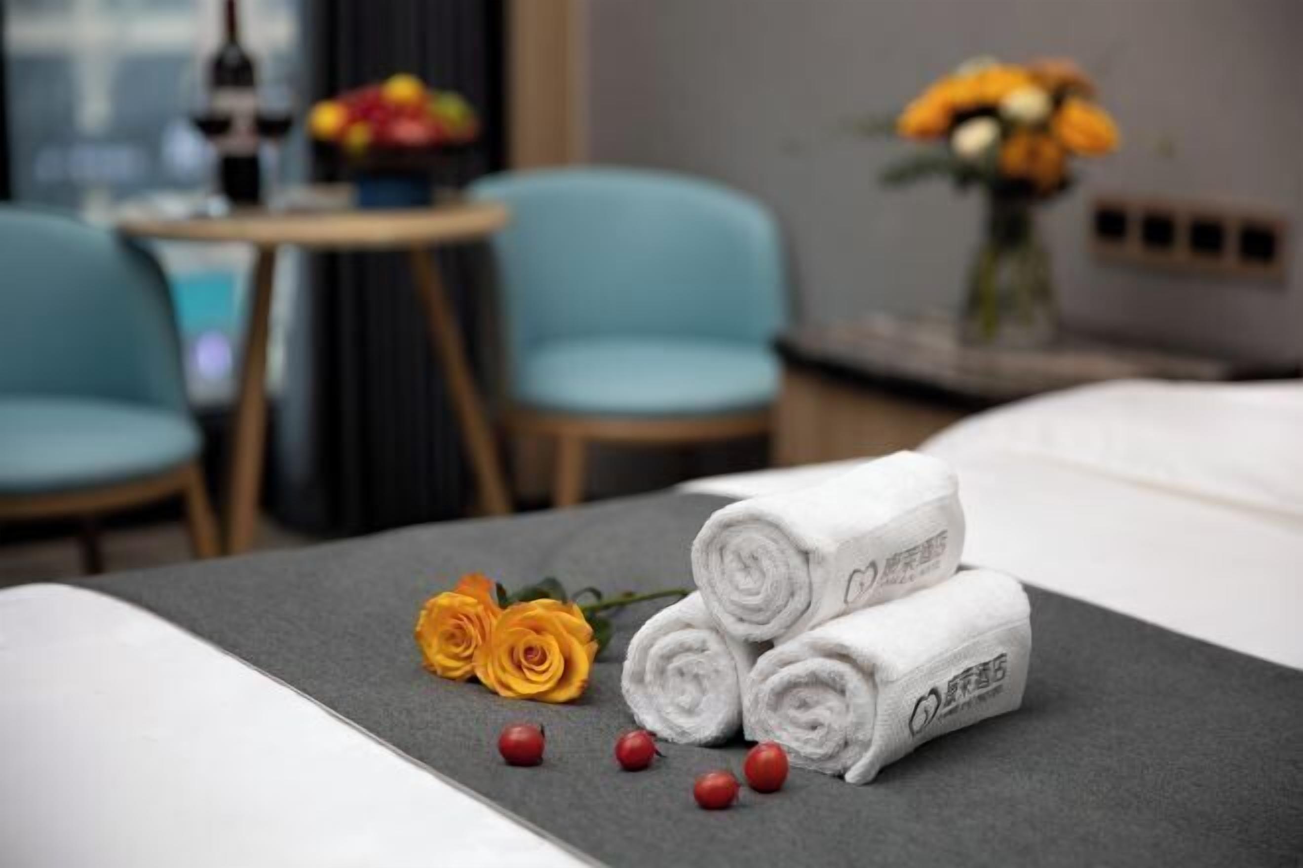 Un lit bien fait, avec des serviettes blanches pliées en rouleau, un vase rempli de fleurs jaunes et une petite table sur laquelle se trouvent une bouteille et des fruits.