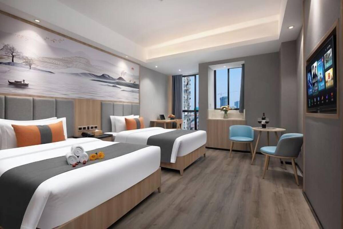 A modern hotel room with two beds, a seating area with chairs, a TV, and a large window offering a city view.