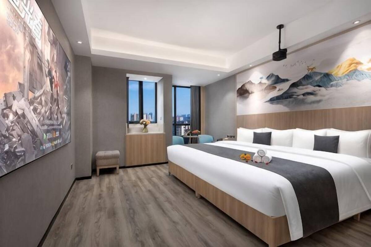A modern hotel room with a large bed, a small desk, a window with a city view, and a mural on the wall.