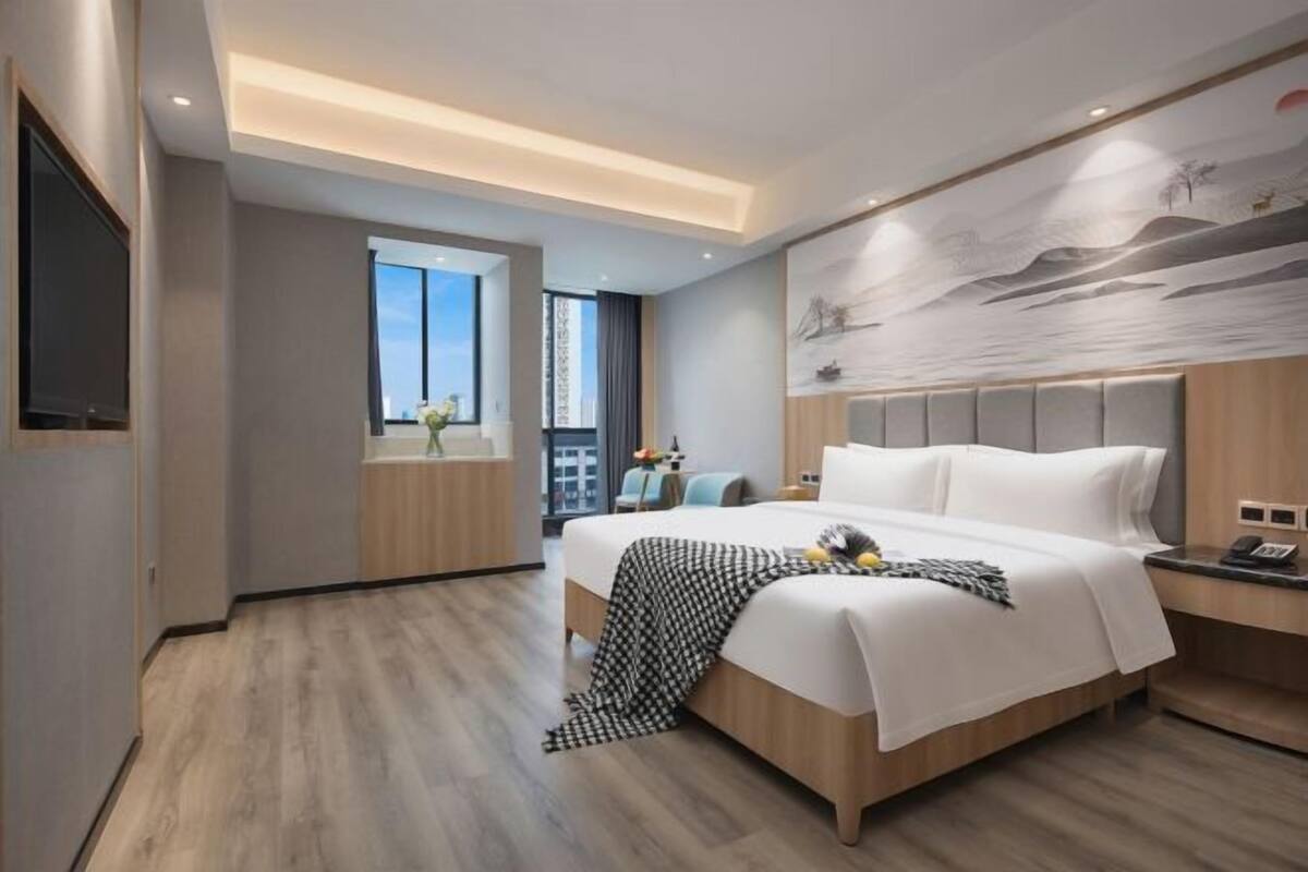 A modern hotel room with a large bed, a flat-screen TV, a window with a city view, and a mural on the wall.