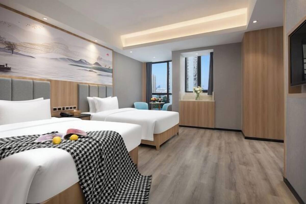 A hotel room with two beds, a small table, a chair, and a window with a view of the city.