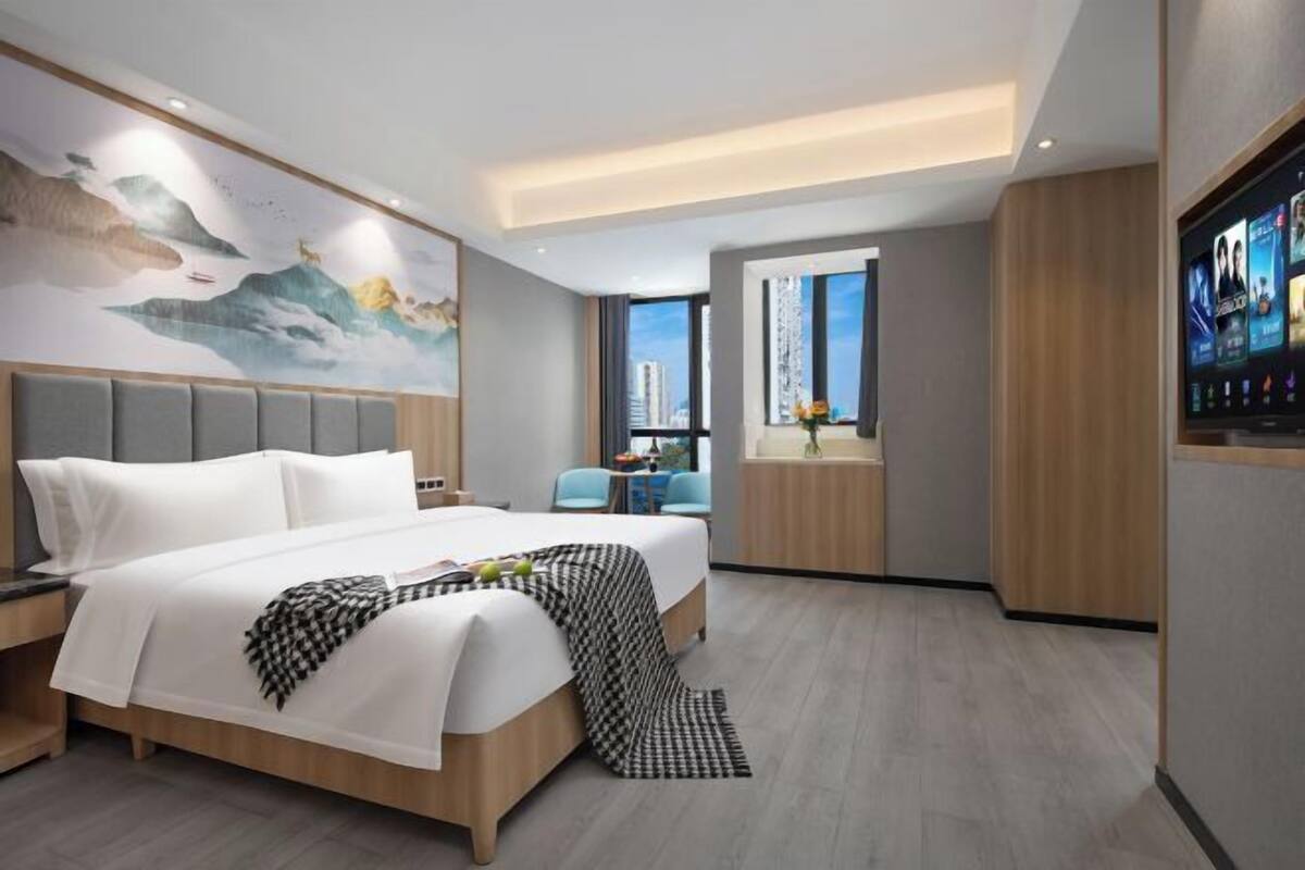 A modern hotel room with a large bed, a city view, a television, and a mural on the wall.