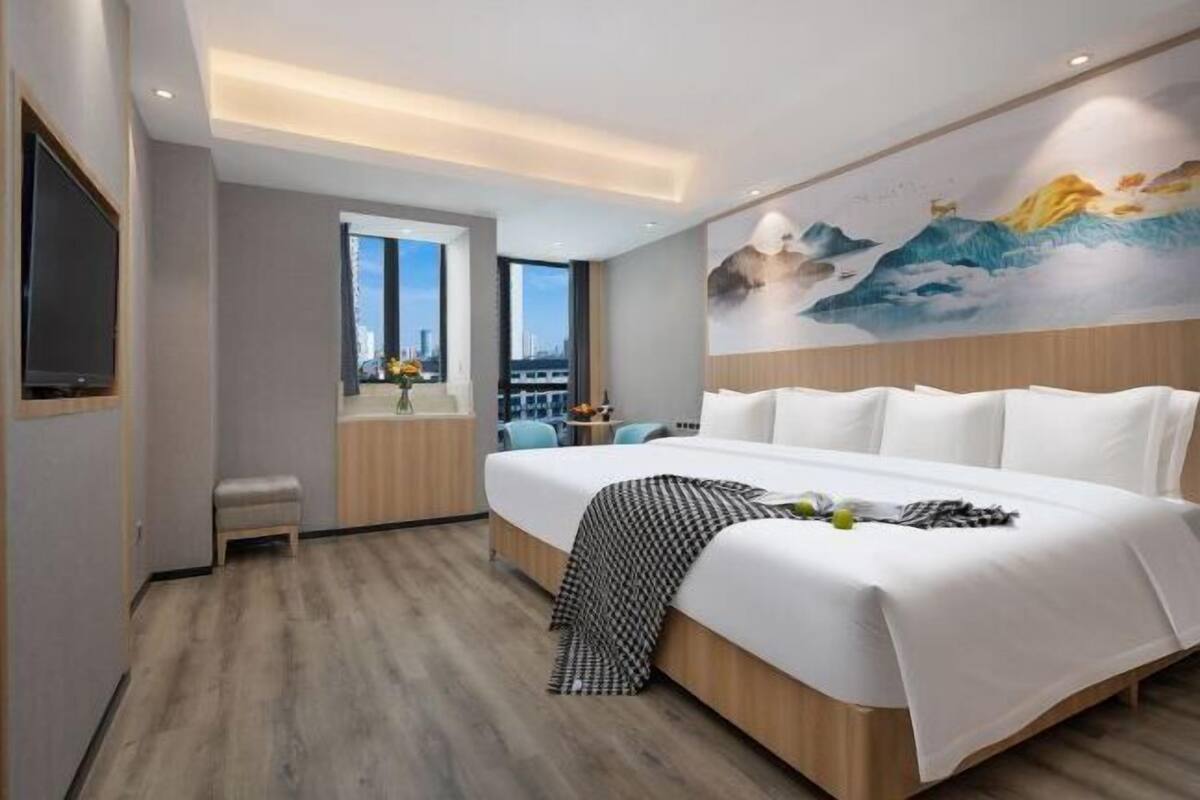 A modern hotel room with a large bed, a television, a window with a city view, and a mural on the wall.