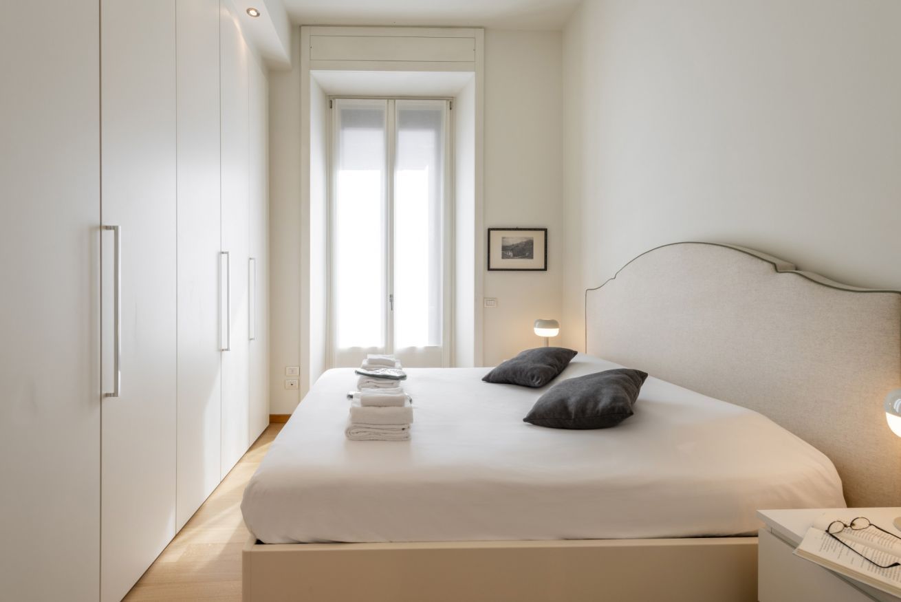 A bedroom with a bed, pillows, a bedside table, a nightstand, a framed picture, and a window.
