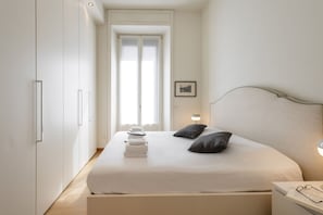 A bedroom with a bed, pillows, a bedside table, a nightstand, a framed picture, and a window.