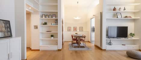 A modern living room with a dining area, built-in shelving, a flat-screen TV, and a large window.