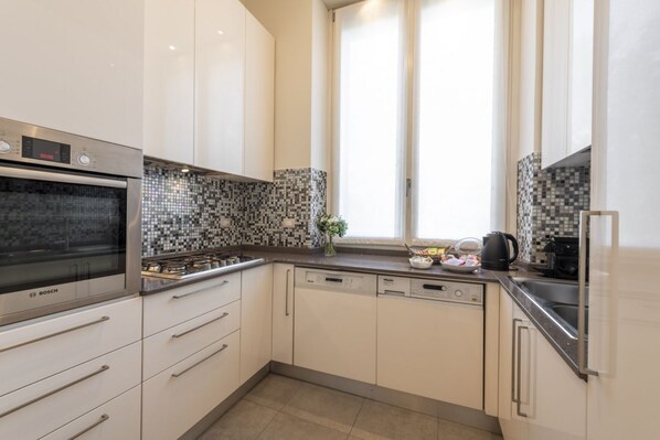 Una cocina moderna con armarios blancos, horno Bosch, lavavajillas y una ventana con cortinas.