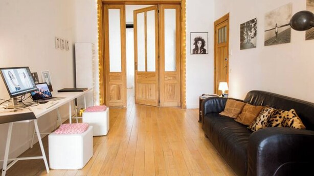 A room with a wooden floor, a black leather sofa, a desk with a computer, and a white chair. There are two white ottomans and a lamp on the sofa. - Beautiful 2-bedroom artist's apartment with all comforts in the heart of Saint-Gilles (Saint-Gilles)