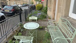 A narrow urban street with parked cars, a green metal bench, a small round table, and a white chair. - Splendid 2bed Apt near Haymarket (Edinburgh)