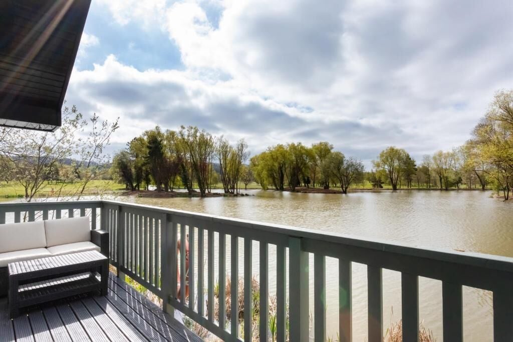 A deck with a sofa and a table, overlooking a calm river with trees and a grassy area in the background.