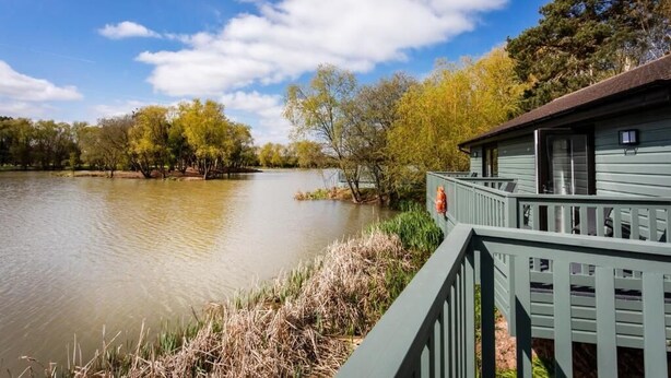 A lakeside cabin with a dock extending into the water, surrounded by trees and a clear sky. - The Vale Golf and Country Club (Pershore)