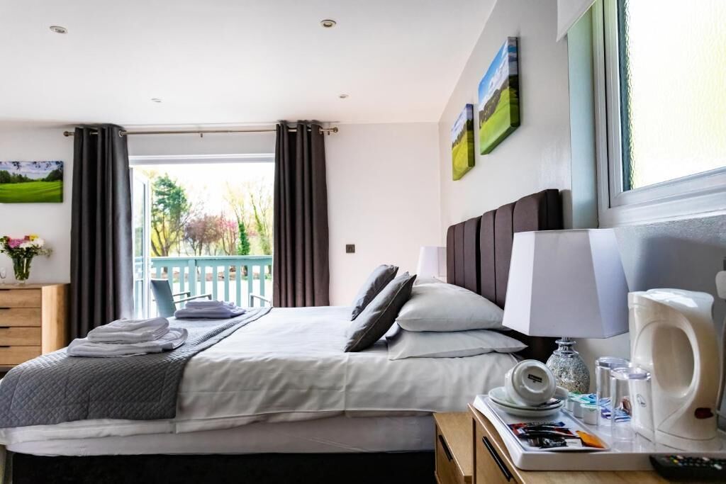 A neatly made bed with pillows, a bedside table with a lamp and a coffee maker, and a view of a balcony with curtains.