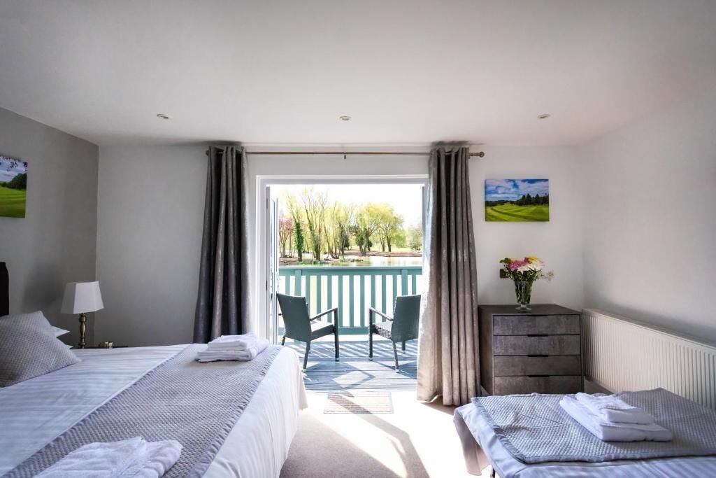 A hotel room with two beds, a balcony with a view of trees and a lake, a dresser, and a lamp.