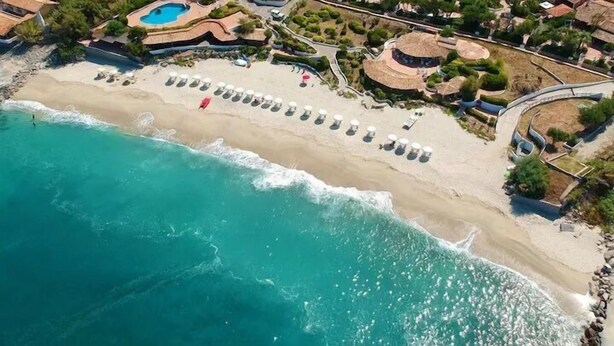 Ett flygbildsfoto av ett strandhotell med en lång strand, en pool och flera byggnader med halmtak.