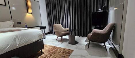 A modern hotel room with a large bed, two armchairs, a small table, and a TV.